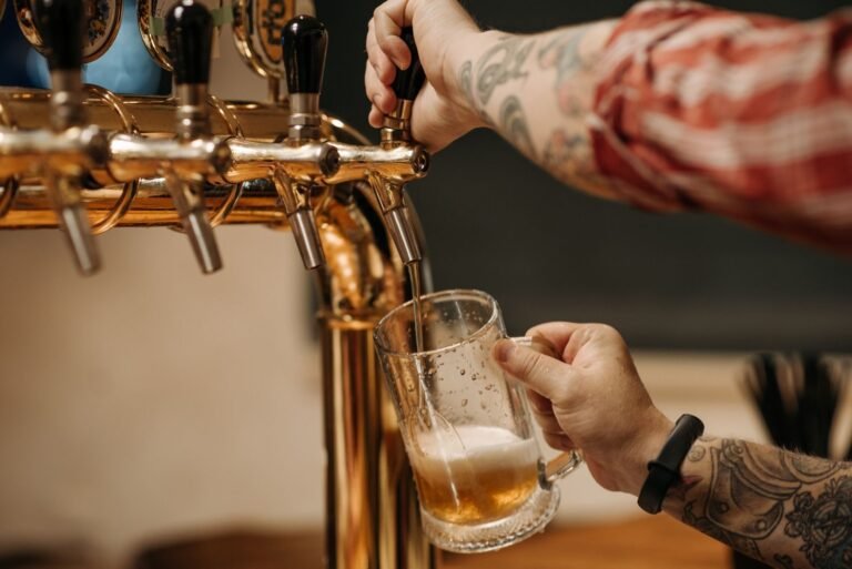 Tattooed bartender pouring a pint of beer from a golden tap, close-up view at a bar.