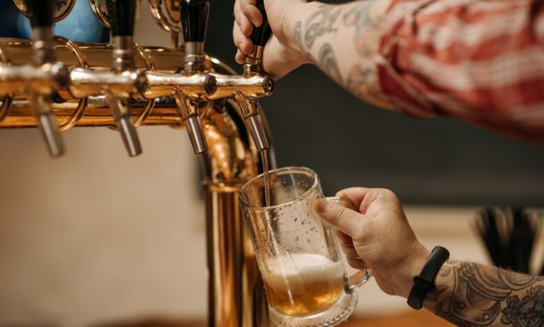 Tattooed bartender pouring a pint of beer from a golden tap, close-up view at a bar.