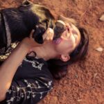 woman in black and white floral dress lying on brown ground with black and brown short dog licking