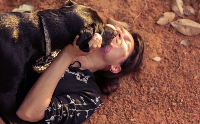 woman in black and white floral dress lying on brown ground with black and brown short dog licking